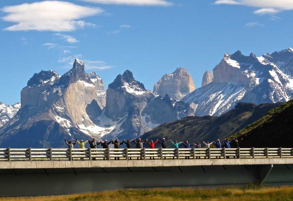 row of hikers with hands linked and raised on a bridge in torres del paine in front of snow-caped jagged mountains