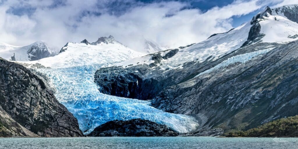 large glacier cascading down rocky snow-capped mountains on a lake on a cloudy day