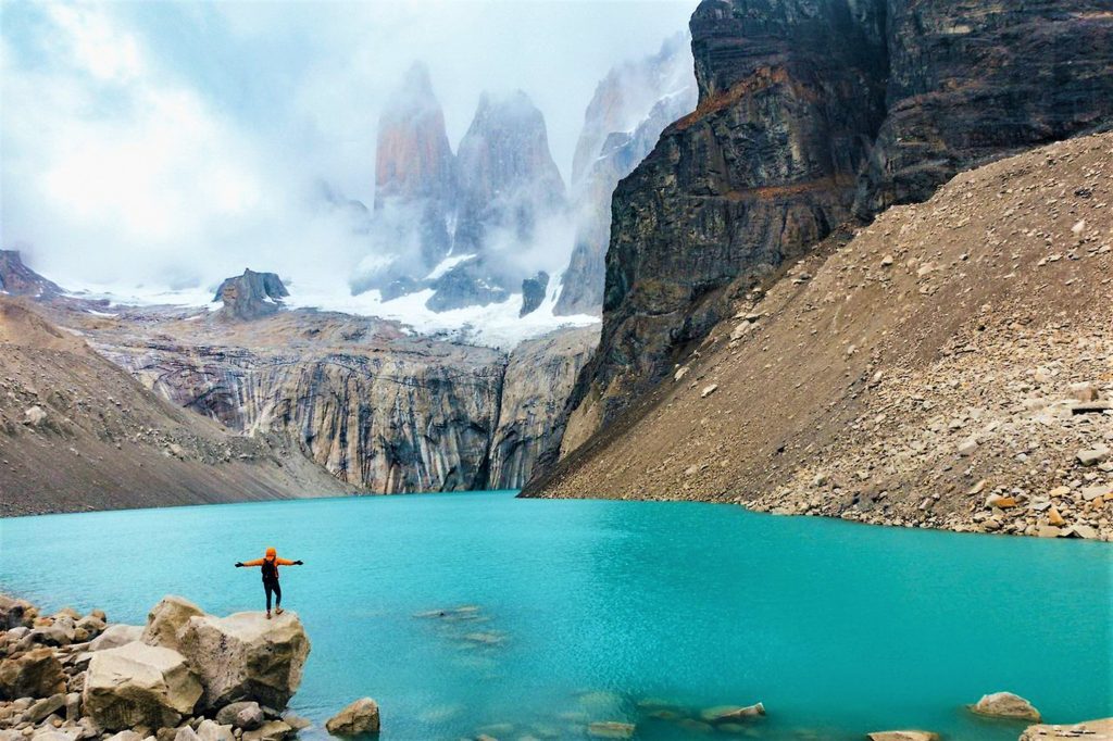 hiker with outstretched arms standing on the edge of a cliff above a glacier lake with granite peaks in the background