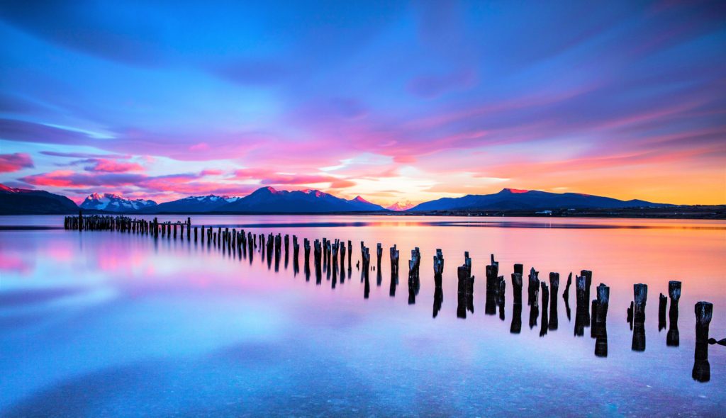 weathered wooden posts remaining from a destroyed pier with a bright sunset behind mountains reflecting on a body of water
