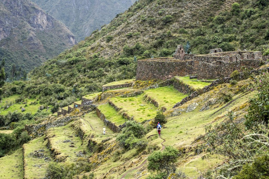 hikers walking on a dirt trail by ancient Incan ruins on grassy terraces near Andes mountains
