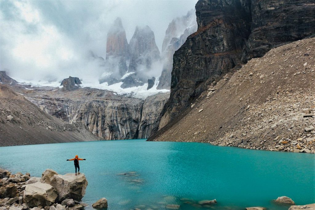 Hiker with outstretched arms on a cliff by a glacial lake and Las Torres granite tower peaks on the W Trek in Patagonia
