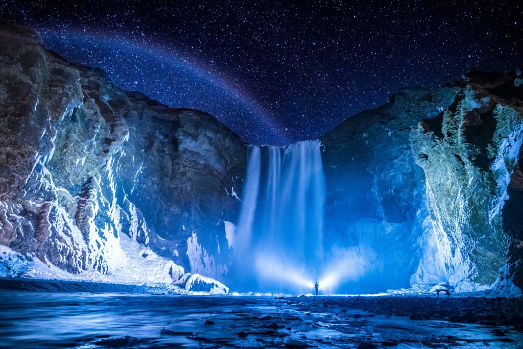 Person standing in front of an icy waterfall surround by snowy mountains on a starry night in Iceland