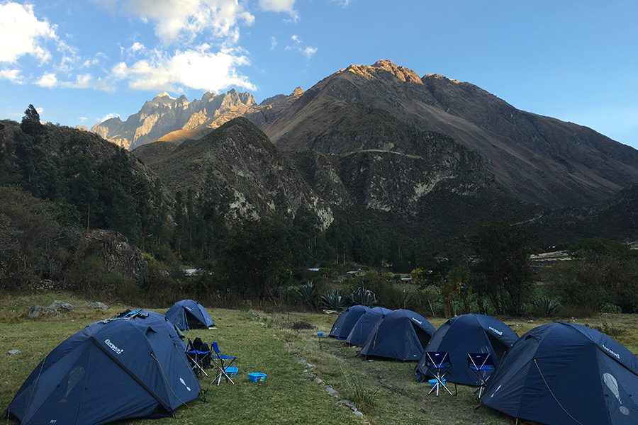 A daytime photograph of blue tents at a trekking campsite in the Sacred Valley, Peru. The background depicts tall mountains in the Andes range