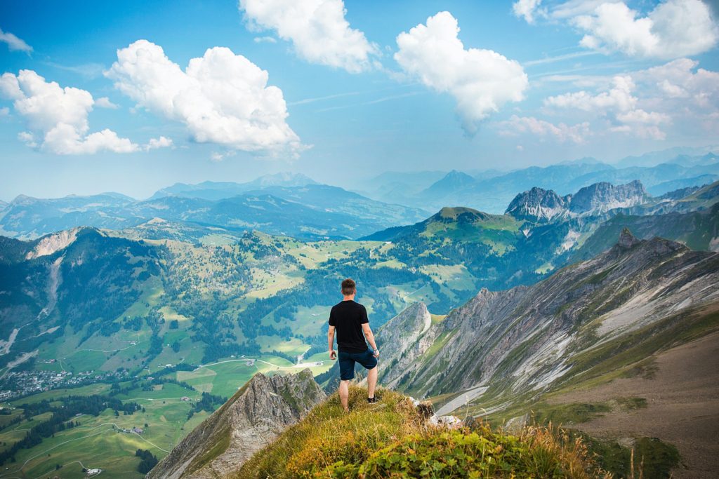 Man standing alone overlooking green mountains with blue skies and clouds above.