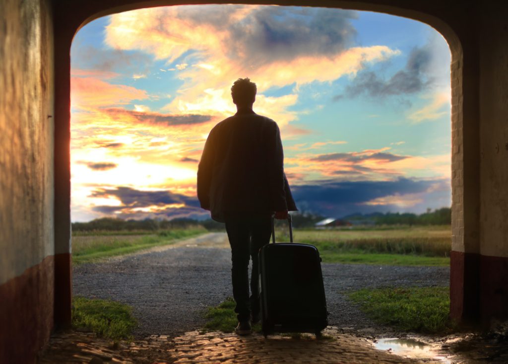 Traveler holding luggage in an archway while looking off into the sunset.