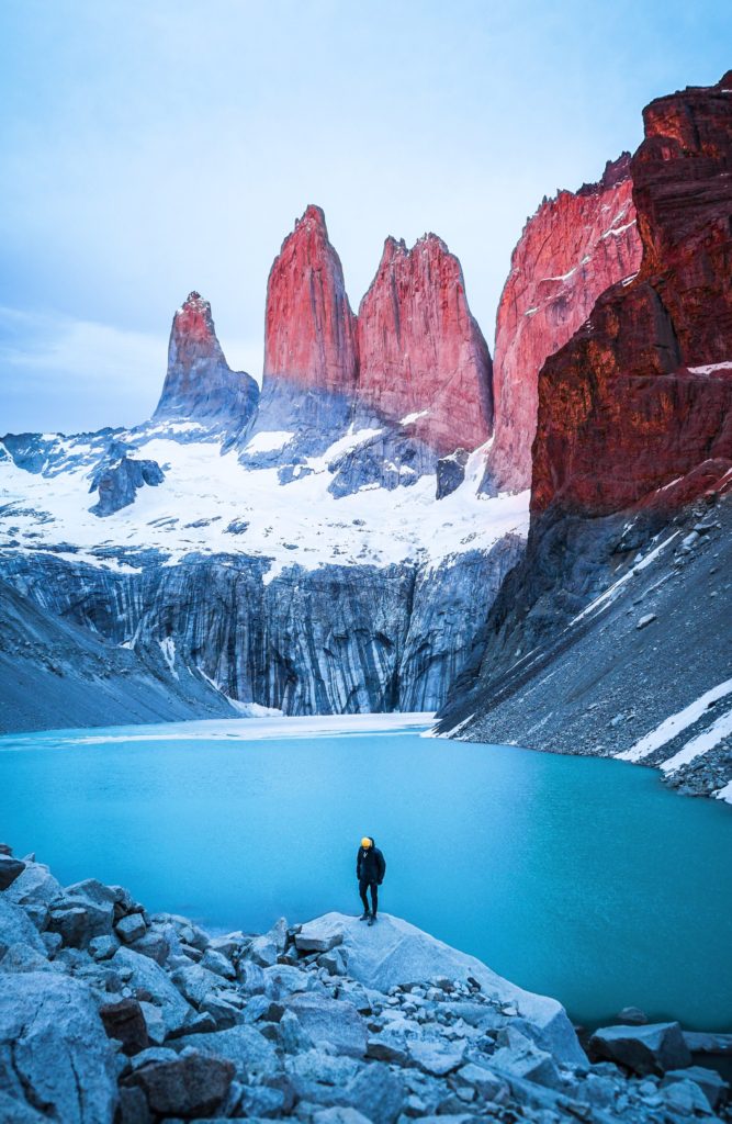 Person standing on a rock in front of blue water and three red granite towers with snowy bases.