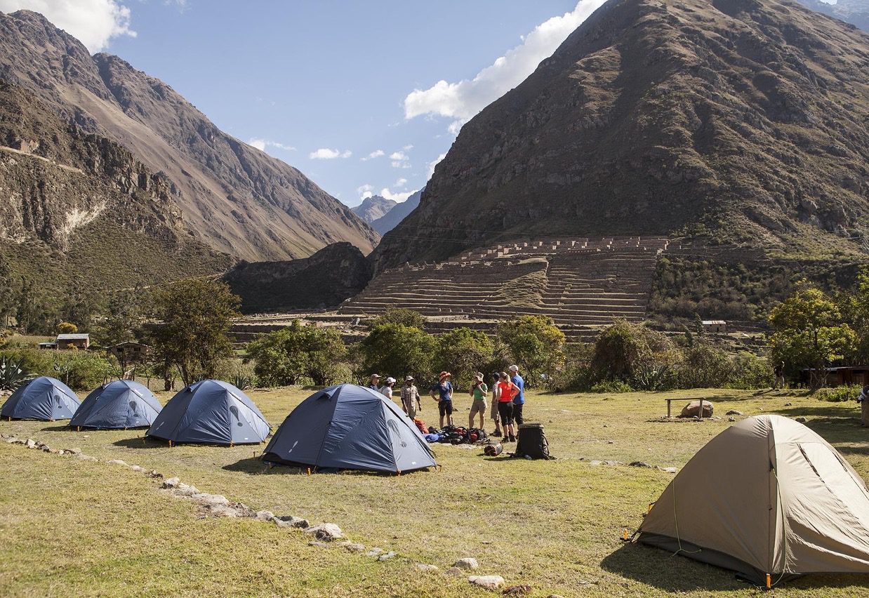 A photograph of several blue tents in a row, for trekkers on an Inca Trail tour to Machu Picchu. The tents are set on flat ground in a grassy valley, and a small group of hikers stands in a circle near them