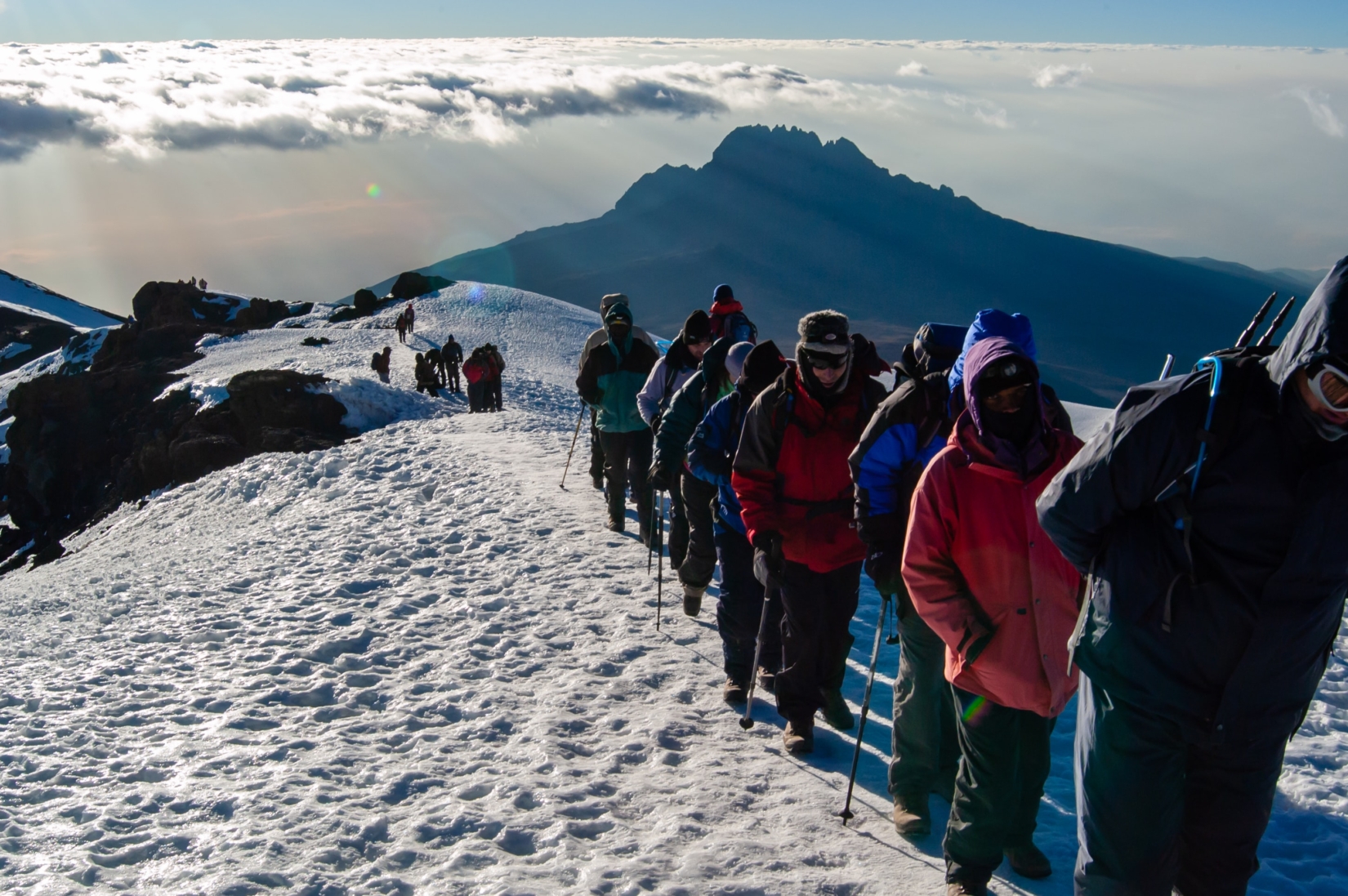 A single line of trekkers climbing a snow-covered mountain above the clouds