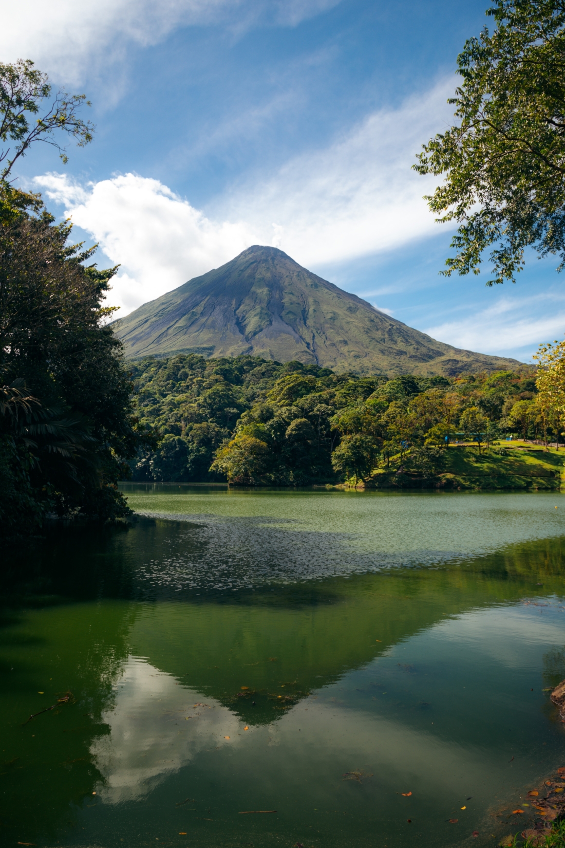 A lake sits at the forefront of the image as focus shifts to the mighty Arenal Volcano emerging from dense jungle