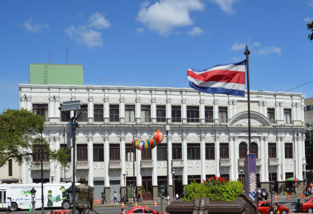 People walking and red taxis passing in front of a white museum building in a city with the Costa Rican flag waving in the wind