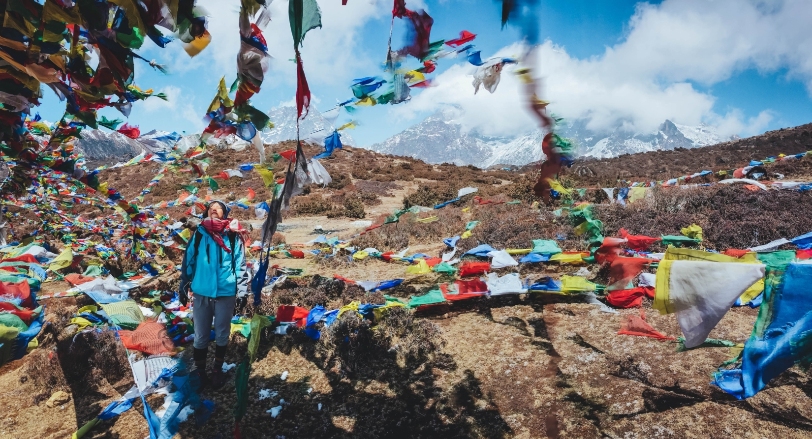 A solo trekker pauses in a sea of prayer flags, blown in the wind, as she treks in the Himalayas