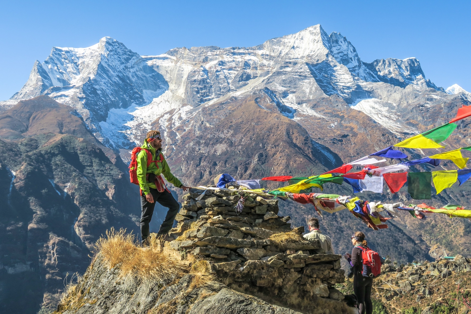 A photograph of a trekker en route to Everest base camp, standing by a rock cairn and Himalayan prayer flags. There are snow-covered mountains in the background