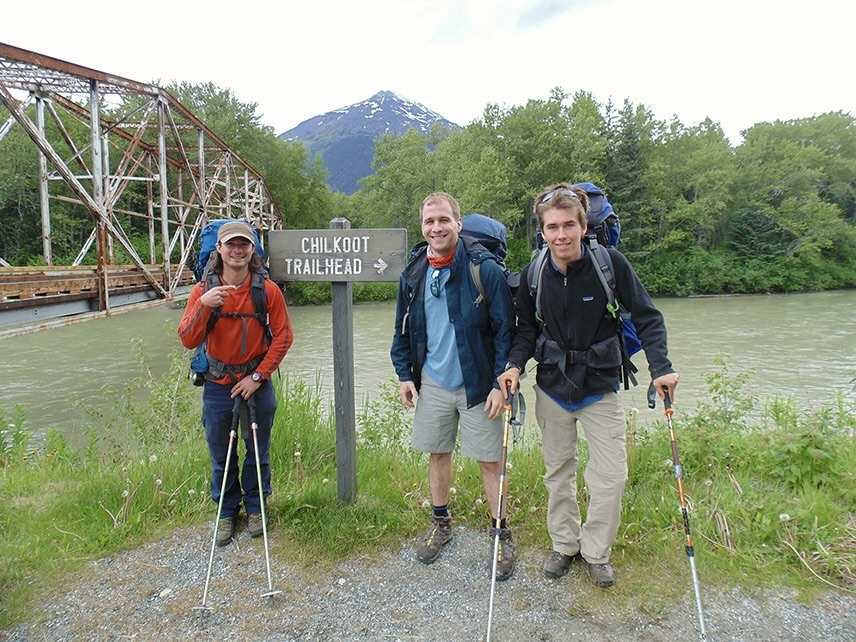 A small group of travelers with hiking poles and gear standing by a lake and rusty bridge at the Chilkoot Trailhead sign.